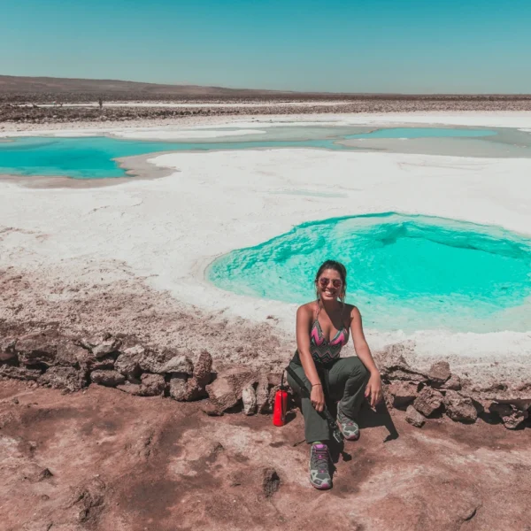 Mulher sentada sorrindo com o cenário das Lagunas escondidas - Atacama ao fundo. Pode-se ver pequenas lagoasde água cristalina e coloração azul turquesa.