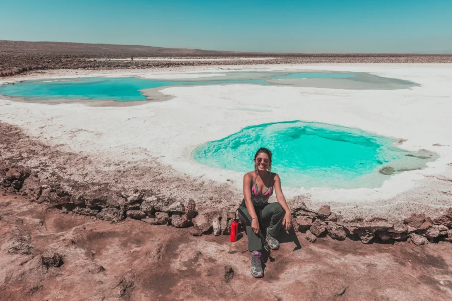 Mulher sentada sorrindo com o cenário das Lagunas escondidas - Atacama ao fundo. Pode-se ver pequenas lagoasde água cristalina e coloração azul turquesa.